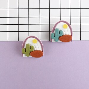 Könnte beinhalten: Zwei violette, weiße und gelbe regenbogenförmige Filzornamente mit einem grünen Kaktus und einem orangefarbenen Bodendesign.