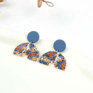 Puede incluir: Un par de pendientes de declaración azules y multicolores con forma de semicírculo. Los pendientes tienen un patrón moteado y un círculo azul en la parte superior.