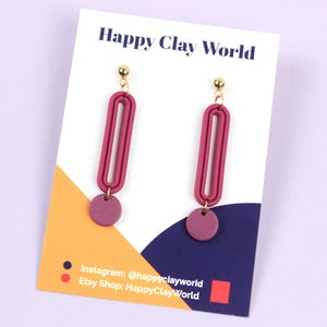 Könnte beinhalten: Ein Paar weinrote Ohrringe mit goldenen Stiften. Jeder Ohrring hat eine längliche, ovale Form mit einem kleinen, kreisförmigen Akzent. Die Ohrringe werden auf einer weißen Karte mit dem Text "Happy Clay World" präsentiert.