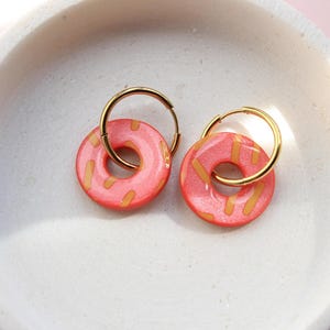 Könnte beinhalten: Ein Paar Creolen mit runden Anhängern in Form von rosa Donuts mit gelben Streuseln. Die Anhänger hängen an dünnen, polierten goldfarbenen Metallreifen. Die Donut-Anhänger haben eine glatte, glänzende Oberfläche und eine kräftige, gesättigte rosa Farbe mit kleinen, rechteckigen gelben Akzenten.