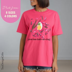 Puede incluir: Camiseta rosa brillante con un diseño temático de comida que incluye un huevo, hierbas y verduras. El texto "[shal-tee-bahr-sh-chay]" está debajo. Disponible en 8 tallas y 4 colores.