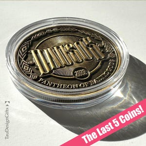May include: A circular bronze-toned metal coin encased in a clear plastic protective capsule. The coin features the embossed text "DIEVS" and "PANTHEON OF BALTIC GODS" with the year "2022" and intricate geometric border patterns.