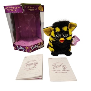 May include: A black and yellow striped electronic Furby toy with green eyes and a yellow beak, accompanied by its original purple and green cardboard display box, an instruction manual, and a Furbish-English dictionary booklet.