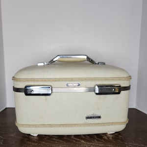 May include: A cream-colored hard-shell train case with a textured exterior. It features a silver-tone metal handle, a horizontal metal band across the front, and two metal latches. The brand name American Tourister is printed on a small badge, and the word Tiara is printed on a separate badge below.
