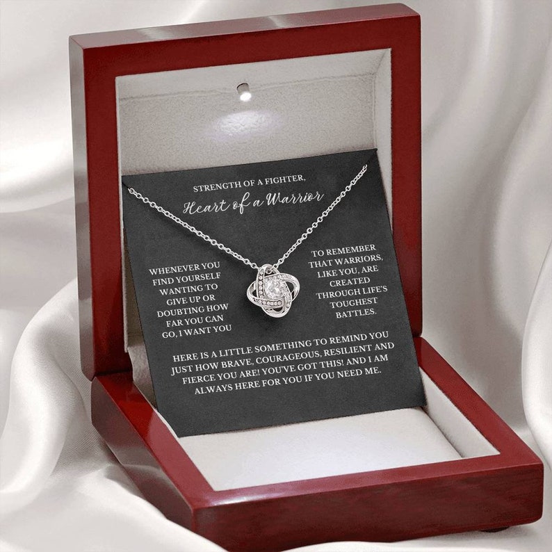 May include: A silver-colored necklace with a knot pendant, presented in a red wooden jewelry box. The box contains a card with the text "Heart of a Warrior" and inspirational messages. The necklace is displayed against a white satin background.