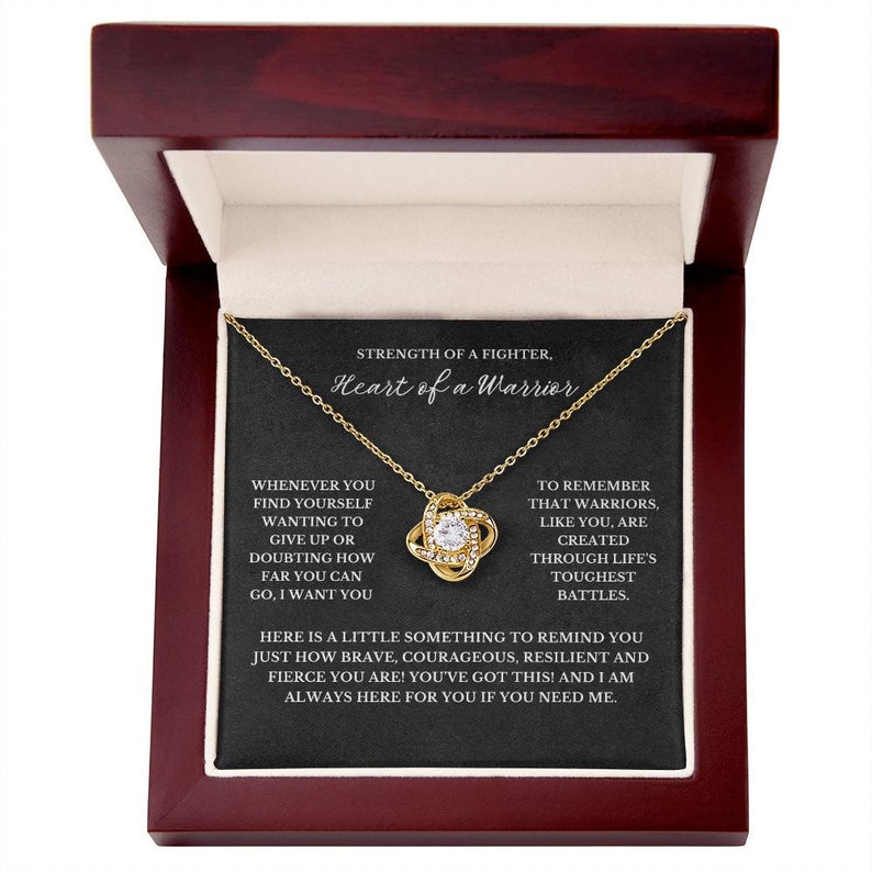 May include: A gold-colored necklace with a knot pendant, presented in a dark wooden box with a black interior. The necklace is displayed on a black card with the text "Strength of a Fighter, Heart of a Warrior".
