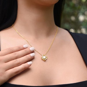 May include: A gold-colored necklace with a pendant featuring a central white stone surrounded by smaller clear stones. The necklace is worn around the neck, with a hand gently touching the chain.