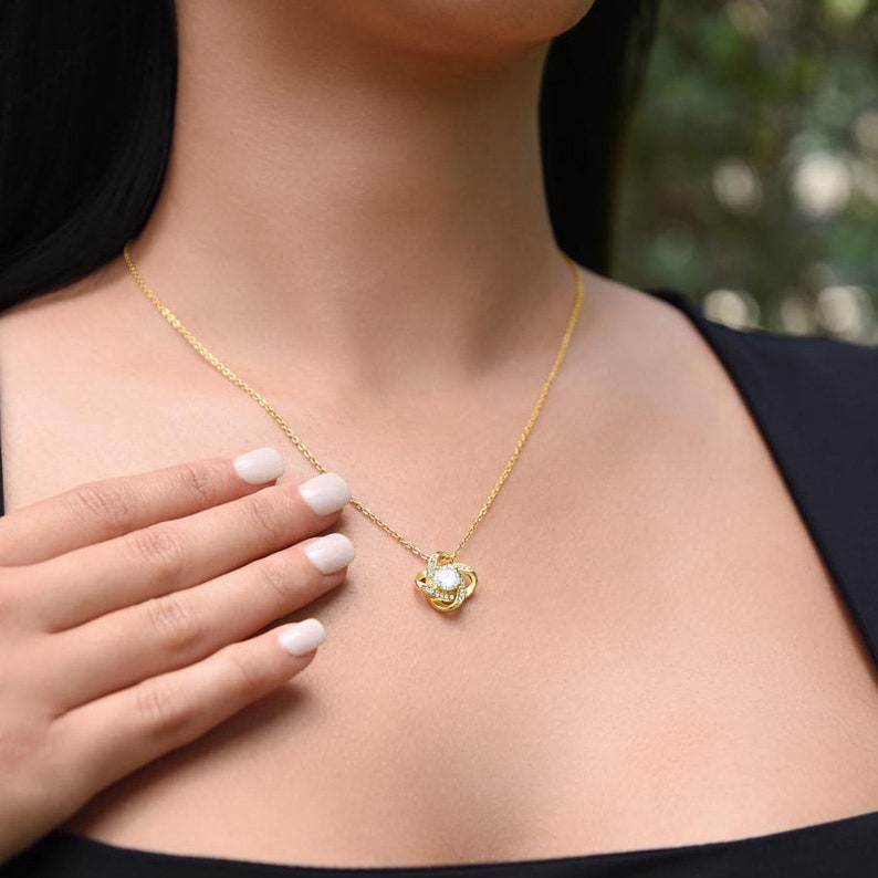May include: A gold-colored necklace with a pendant featuring a central white stone surrounded by smaller clear stones. The necklace is worn around the neck, with a hand gently touching the chain.