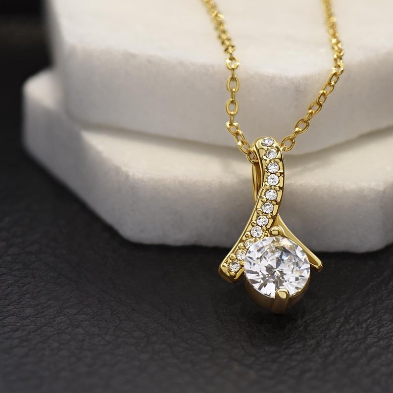 May include: A gold necklace with a pendant featuring a large, clear, round gemstone surrounded by smaller, clear gemstones. The pendant is shaped like a teardrop and has a delicate, curved design.