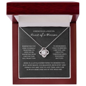 May include: A silver-toned necklace with a knot pendant, presented in a dark red jewelry box. The pendant features a central clear stone surrounded by smaller stones. The box has a black interior with inspirational text.