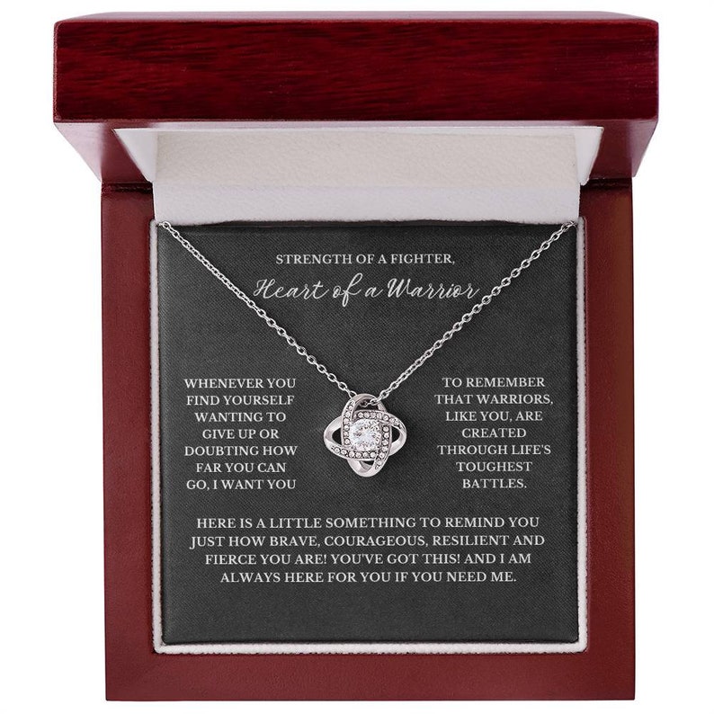 May include: A silver-toned necklace with a knot pendant, presented in a dark red jewelry box. The pendant features a central clear stone surrounded by smaller stones. The box has a black interior with inspirational text.