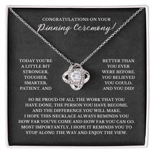 May include: A silver necklace with a crystal knot pendant in a black gift box. The box has a message that reads "Congratulations on your Pinning Ceremony!" and "Today you're a little bit stronger, tougher, smarter, patient, and better than you ever were before. You believed you could, and you did!"