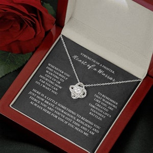 May include: A silver-colored necklace with a knot pendant, presented in a red jewelry box. The box contains a card with the text "Heart of a Warrior" and inspirational quotes. A red rose is visible in the background.