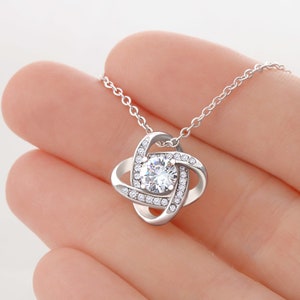 May include: A silver necklace with a knot pendant featuring a clear crystal in the center. The knot is surrounded by smaller clear crystals.