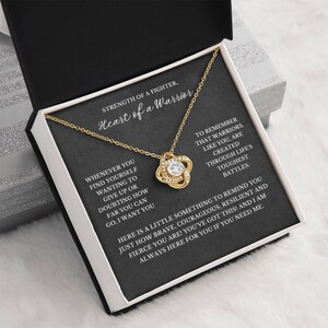 May include: A gold-colored necklace with a knot pendant, set in a black box with white accents. The box contains a card with the text "Strength of a Fighter, Heart of a Warrior." The necklace is displayed on a white surface.