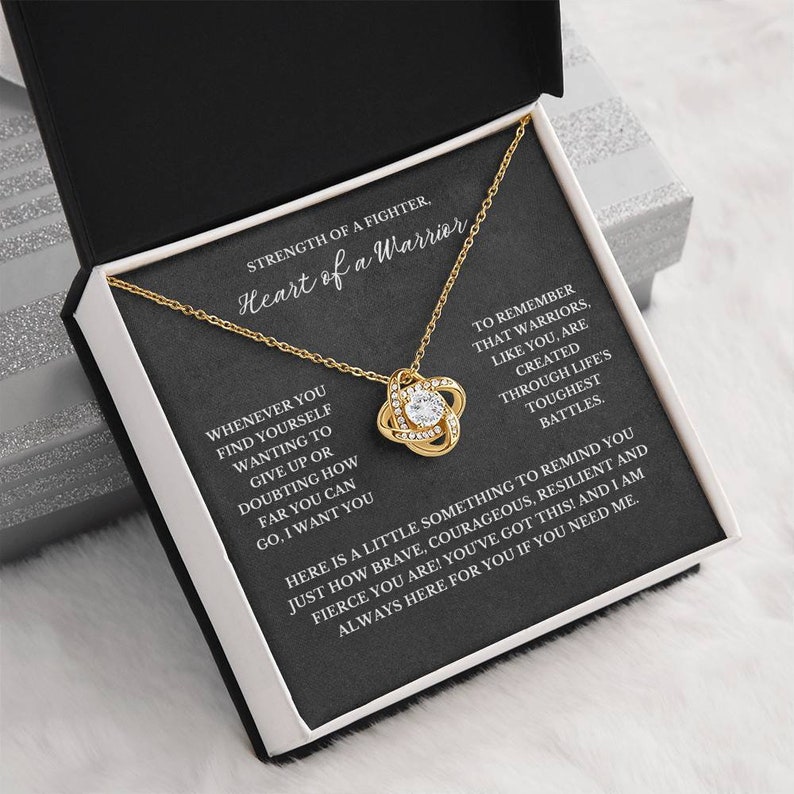 May include: A gold-colored necklace with a knot pendant, set in a black box with white accents. The box contains a card with the text "Strength of a Fighter, Heart of a Warrior." The necklace is displayed on a white surface.