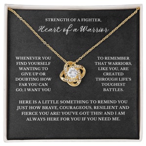 May include: A gold-colored necklace with a pendant featuring a central clear crystal surrounded by smaller crystals. The necklace is displayed on a black background with white text that reads "STRENGTH OF A FIGHTER, Heart of a Warrior."