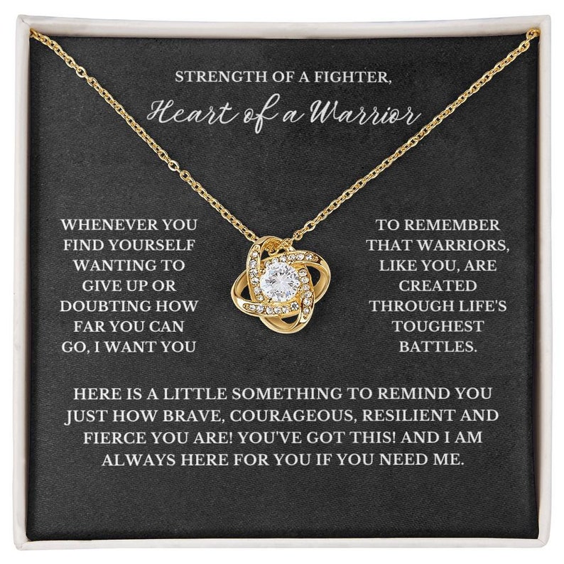 May include: A gold-colored necklace with a pendant featuring a central clear crystal surrounded by smaller crystals. The necklace is displayed on a black background with white text that reads "STRENGTH OF A FIGHTER, Heart of a Warrior."