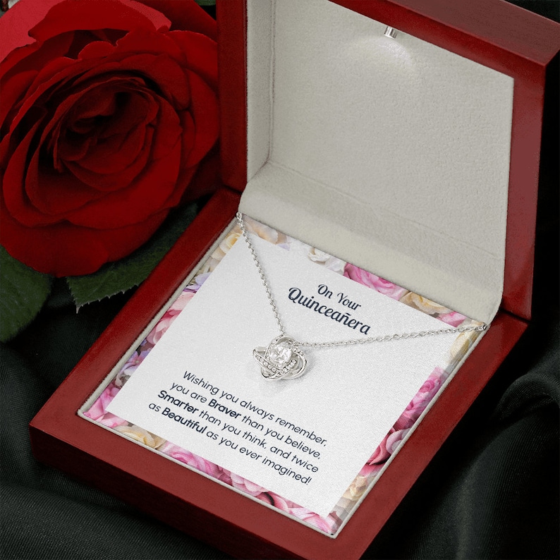 May include: A red velvet jewelry box with a silver necklace inside. The necklace has a knot pendant with a clear crystal in the center. The box has a card inside that reads "On Your Quincea&ntilde;era Wishing you always remember you are Braver than you believe. Smarter than you think, and twice as Beautiful as you ever imagined!"