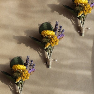 May include: Three small bouquets of dried flowers, each tied with twine and secured with a pin. The bouquets feature yellow flowers, purple lavender, and yellow foliage.