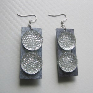 Puede incluir: Un par de pendientes plateados con dos discos redondos, texturados, plateados en cada pendiente. Los pendientes están unidos a una base rectangular de madera gris.