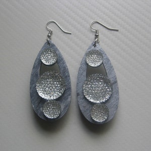 Puede incluir: Un par de pendientes colgantes plateados con tres piedras redondas, brillantes, de color plateado. Los pendientes están hechos de un material gris y tienen forma de lágrima.