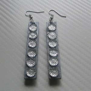 Puede incluir: Un par de pendientes de brillo plateado con forma rectangular y seis acentos redondos de brillo plateado en cada pendiente.