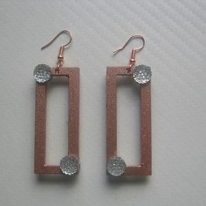 Puede incluir: Un par de pendientes rectangulares de color cobre con un acabado texturizado. Cada pendiente presenta una pequeña bola brillante de color plateado en la parte superior e inferior. Los pendientes tienen ganchos para las orejas de color cobre.