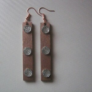 Puede incluir: Un par de pendientes colgantes con colgantes rectangulares de madera, cada uno con tres adornos circulares plateados. Los pendientes tienen ganchos para las orejas de color cobre. Diseño sencillo y elegante.