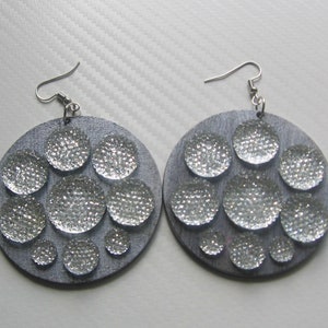 Puede incluir: Un par de pendientes de madera redonda con purpurina plateada y varios círculos de purpurina plateada.