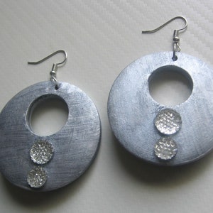 Puede incluir: Un par de pendientes de plata con un diseño circular grande y dos pequeños diamantes de imitación plateados. Los pendientes tienen un cierre de gancho simple.