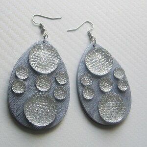 Puede incluir: Un par de pendientes de plata en forma de lágrima con fondo gris y varios círculos plateados con purpurina.