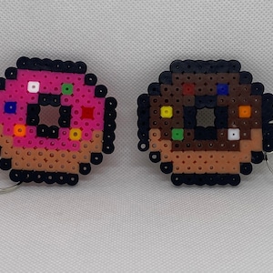 May include: Two pixel art donut keyrings. One has pink icing with colourful sprinkles, the other has chocolate icing with colourful sprinkles. Both have a brown base and black outline. Each keyring has a silver ring.