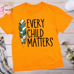 May include: Orange t-shirt with a black and white feather design and the text "Every Child Matters".