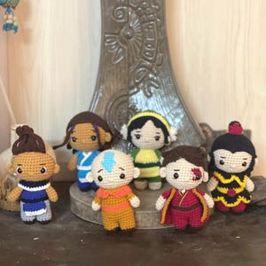 May include: A set of six crocheted figures, each representing a character from the animated series Avatar: The Last Airbender. The figures are all standing on a wooden surface. The characters include Aang, Katara, Sokka, Toph, Zuko, and Azula.