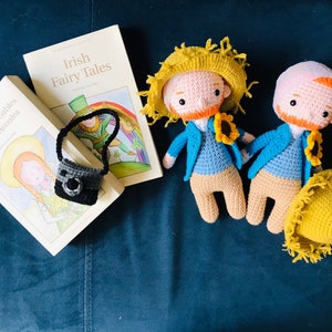 Könnte beinhalten: Zwei gehäkelte Puppen mit blauen Hemden, braunen Hosen und Sonnenblumen-Akzenten. Die Puppen sitzen auf einer blauen Oberfläche mit zwei Büchern, eines mit dem Titel "Irish Fairy Tales" und das andere mit dem Titel "Anne of Green Gables & Anne of Avonlea".