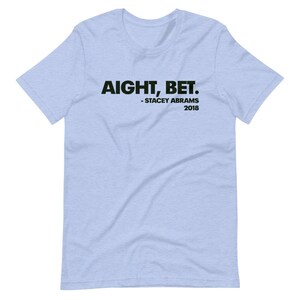 May include: Light blue t-shirt with the text "AIGHT, BET." in black, followed by "Stacey Abrams 2018" in smaller black text.