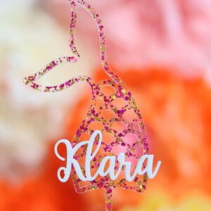Op de afbeelding: Een roze en gouden glitter-zeemeerminstaart caketopper met de naam "Klara" erop.