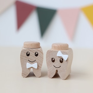 Op de afbeelding: Twee houten doosjes in de vorm van tanden met geschilderde gezichten. Het doosje links heeft een witte strik en het doosje rechts heeft een witte strik. Beide doosjes hebben zwarte ogen en een glimlach.