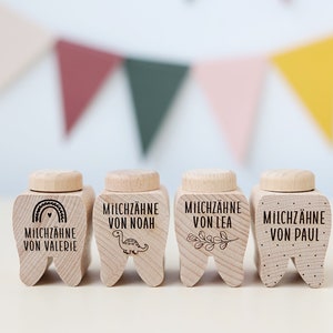 Op de afbeelding: Vier houten tandendoosjes met verschillende ontwerpen. De doosjes zijn gevormd als tanden en hebben de tekst "Milchzähne von" gevolgd door een naam. De ontwerpen omvatten een regenboog, een dinosaurus, een tak en stippen.