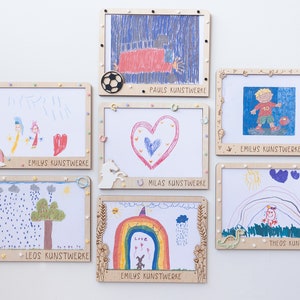 Könnte beinhalten: Eine Sammlung gerahmter Kinderkunstwerke, die an einer weißen Wand ausgestellt sind. Die Rahmen sind aus Holz und haben eine hellbraune Farbe. Jeder Rahmen hat ein anderes Design und enthält den Namen des Kindes und die Worte "Kunstwerke". Die Kunstwerke beinhalten Zeichnungen eines Herzens, eines Regenbogens, eines Fußballspielers, eines Baumes und eines Einhorns.