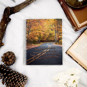 Puede incluir: Un cuaderno de espiral con una cita motivacional sobre dar pasos en la dirección correcta. La portada presenta una fotografía de un camino sinuoso a través de un bosque con hojas de otoño coloridas.