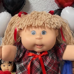 May include: A doll with blonde yarn hair tied with red ribbons, wearing a red and black plaid dress with a red bow. The doll has blue eyes and is surrounded by a red teddy bear, a black teddy bear, other small dolls, and heart-shaped balloons.
