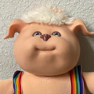 Puede incluir: Un juguete de peluche con cara rosa, pelaje blanco y ojos azules. El juguete lleva un mono amarillo con tirantes arcoíris y un pequeño caballo bordado en la parte delantera.