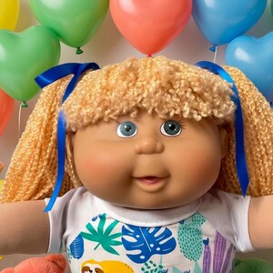 May include: A doll with blonde yarn hair tied with blue ribbons, wearing a white shirt featuring a colorful sloth and tropical leaf print, white lace-trimmed shorts, and blue shoes, surrounded by small baby dolls, teddy bears, and candy.