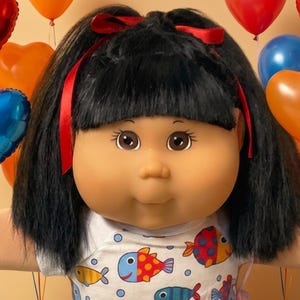 May include: A doll with black hair and red ribbons wearing a white shirt with a colorful fish pattern. The doll has tan skin and is surrounded by small plush bears, plates of brownies and cupcakes, and various heart-shaped balloons.