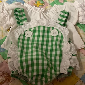 Op de afbeelding: Een groene en witte gingham baby romper met witte kantversiering en twee witte knopen. De romper wordt gecombineerd met een wit geruit topje. De outfit ligt op een groen en geel patchwork dekbed.