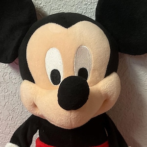 May include: A plush Mickey Mouse doll with black ears, a black nose, and a cream-colored face. The doll wears a red pair of shorts with two white buttons and white gloves. Two yellow plush balls are in front of the doll.