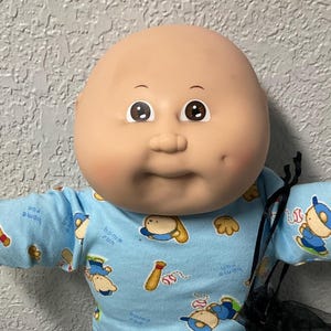 May include: A Cabbage Patch Kids doll with a bald head, wearing a light blue baseball-themed shirt and black pants. The doll has white shoes and is posed with arms outstretched. A black mesh bag is visible.