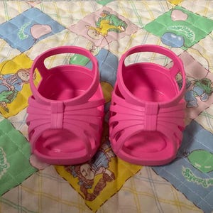 May include: A pair of bright pink plastic sandals. The sandals have a cage-like design with a strap around the ankle and a rounded toe. They are sitting on a colorful patchwork quilt with various cartoon characters.